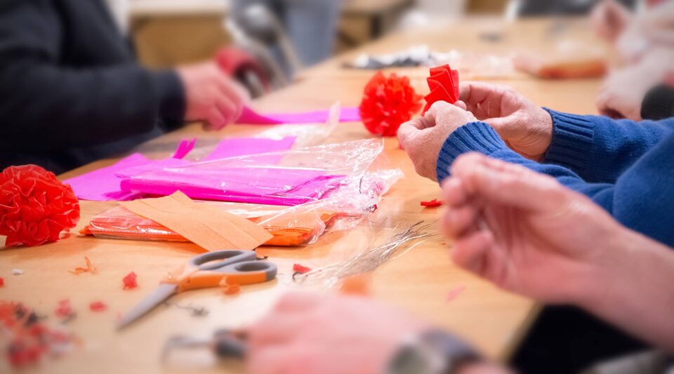 Horizontal composition photography in letterbox image technique, of multiple unrecognizable person, volunteers manufacture and produce red, magenta and orange colored crepe paper decorations, homemade and craft activity. Selective focus on a pair of hands of an elderly person in background with a blue pullover. There are several tools and paper scraps on the table.