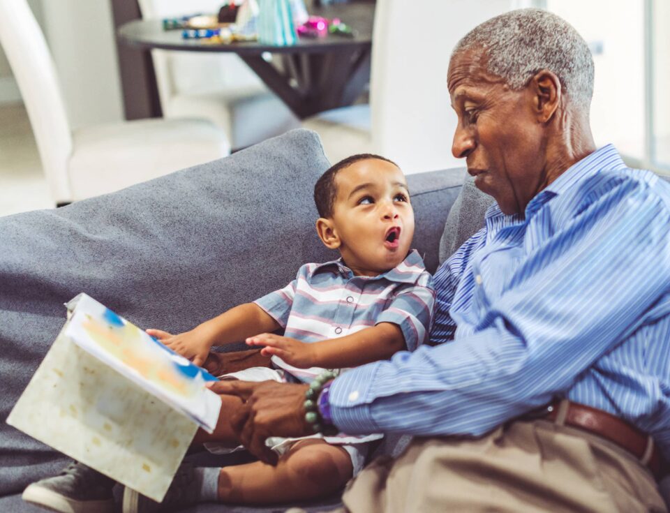 Adorable toddler with his grandfather relaxing at home reading a book together